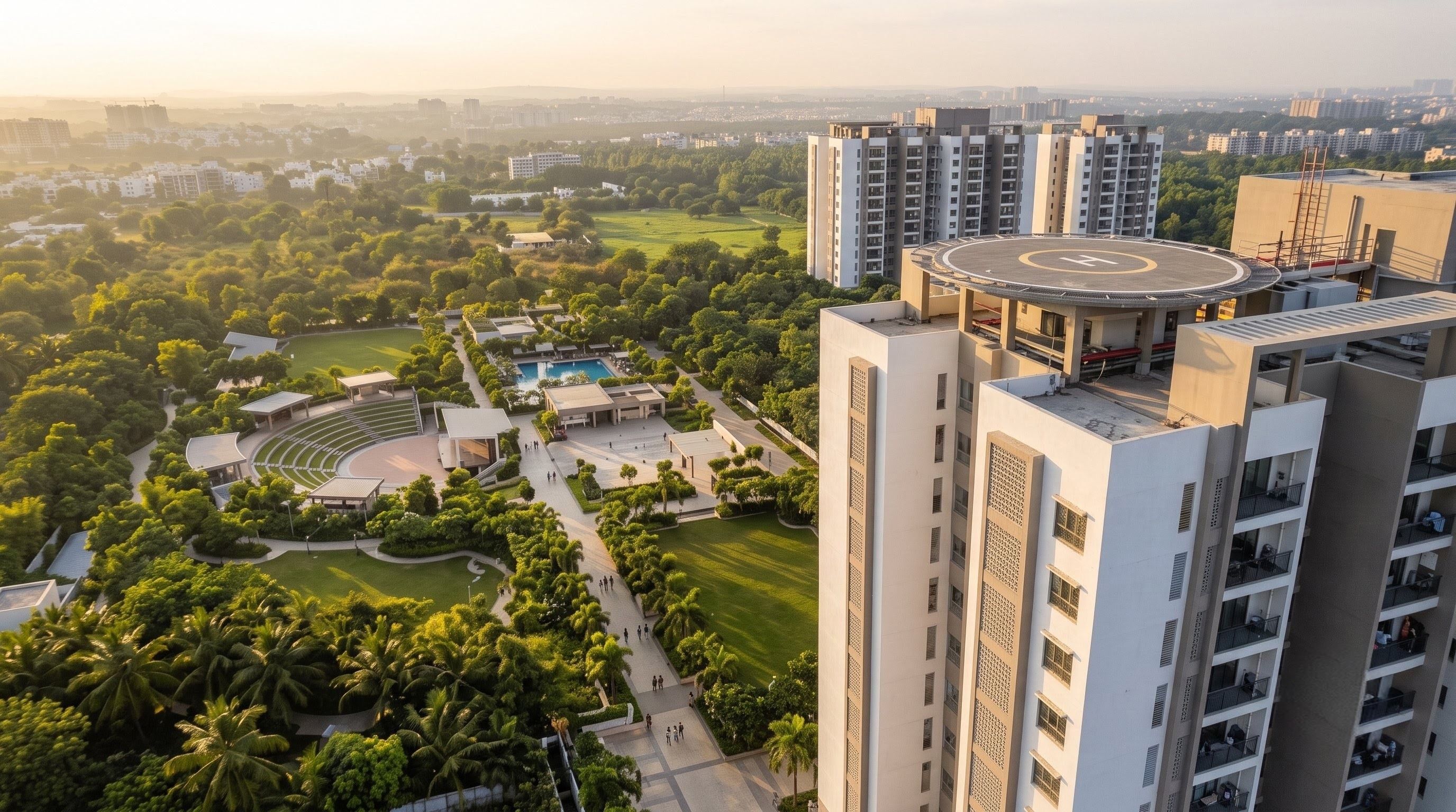 Residential towers at sunrise, seen from the air, beside a wide park with lawns, walkways, an open-air theatre and a pool.